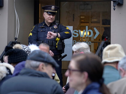 A Wellesley, Mass., police officer glances at his watch as customers and bystanders form a line outside a Silicon Valley Bank branch location, Monday, March 13, 2023, while waiting for the branch to open, in Wellesley, Mass. (AP Photo/Steven Senne)